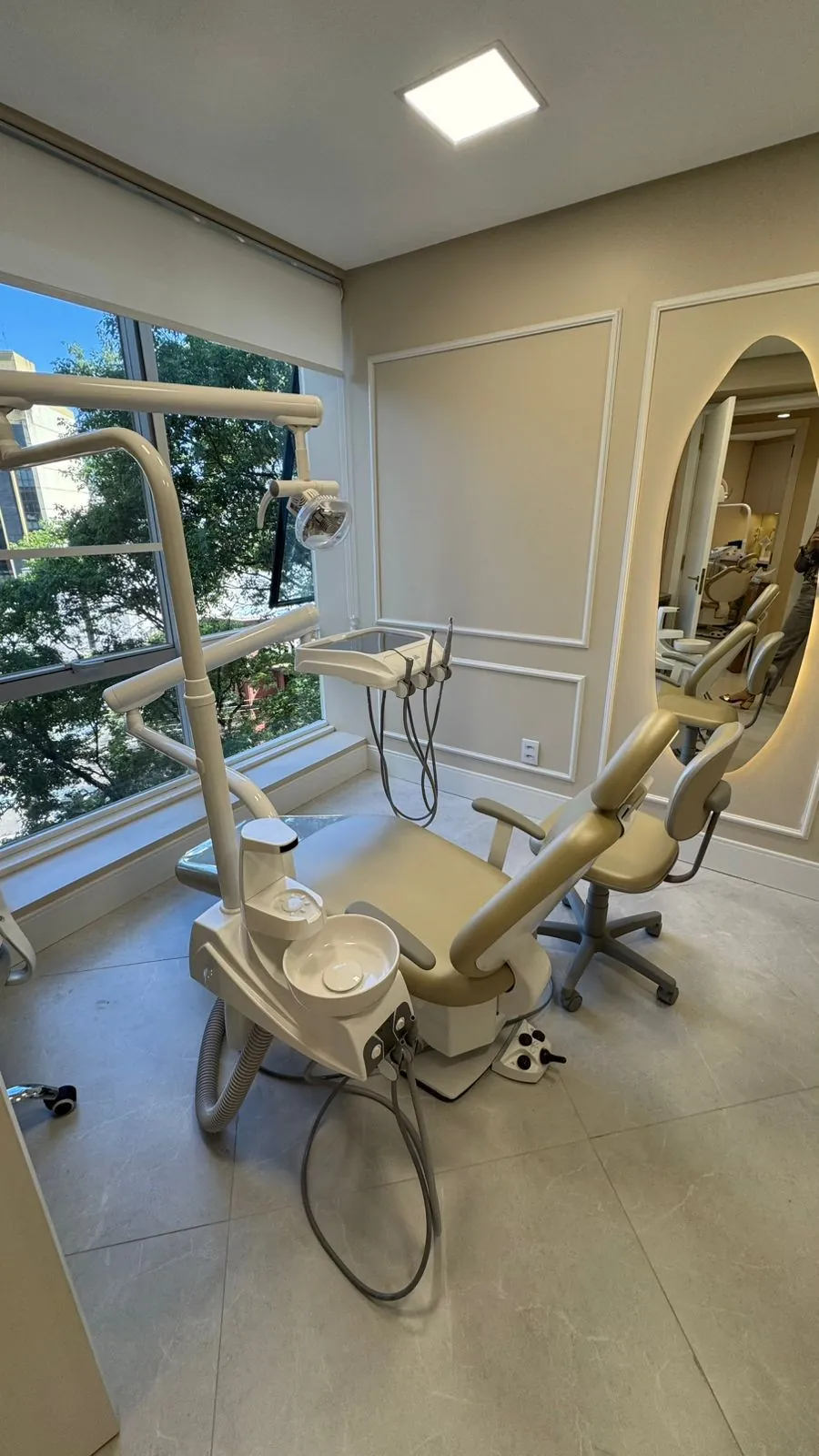 Sala de atendimento com cadeira odontológica bege, janela ampla e espelho iluminado